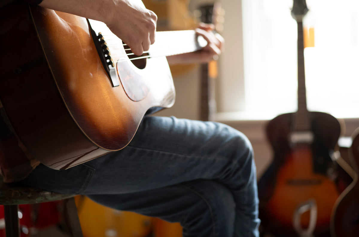 Person holding an acoustic guitar in a room with other guitars in the background