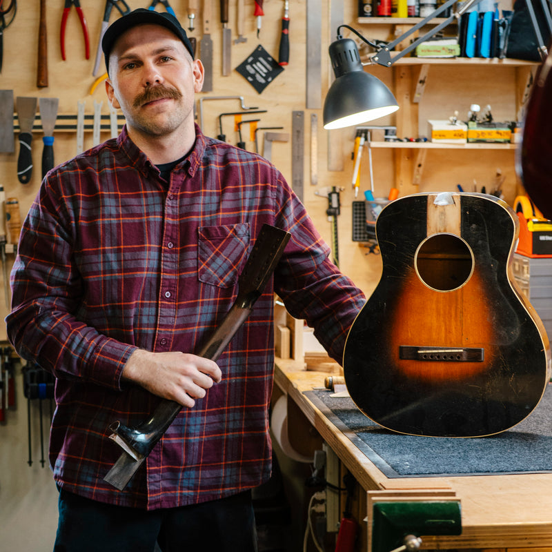 Man holding a vintage Kalamazoo guitar in a workshop with tools and equipment in the background