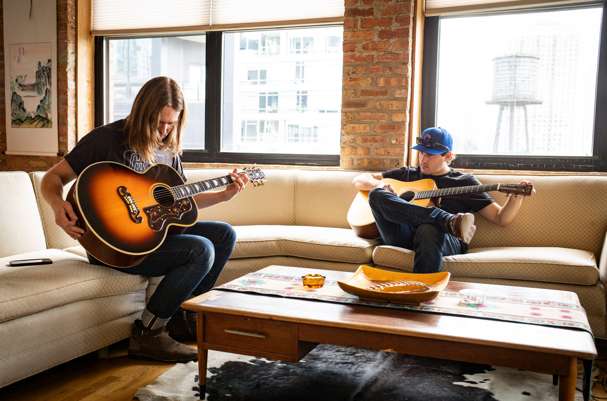 Two people playing vintage guitars on a couch in a living room with large windows.