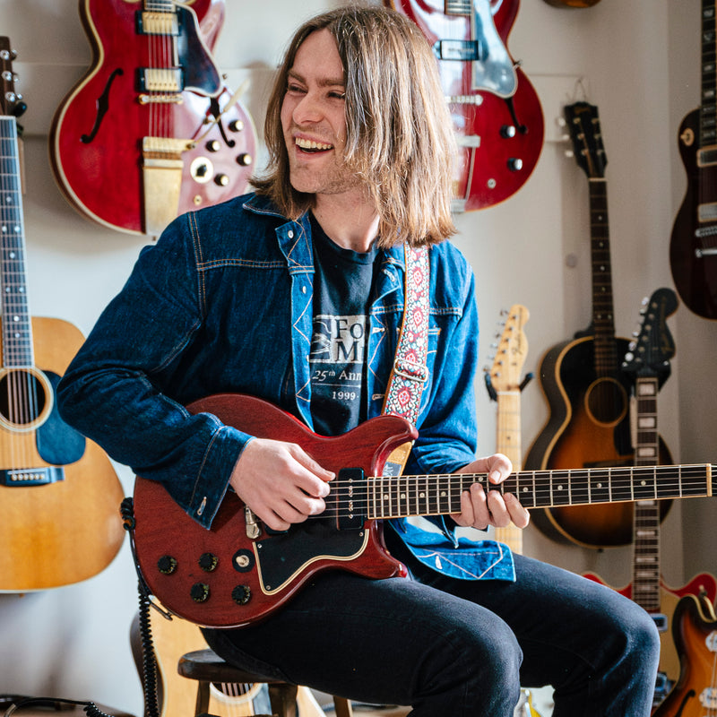 Man playing a vintage Gibson electric guitar with guitars on the wall in the background.
